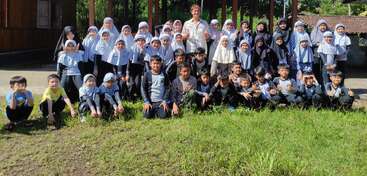 A large group of school children and their teacher pose outdoors for a photo. Most children wear uniforms and headscarves. Everyone looks cheerful and happy.
