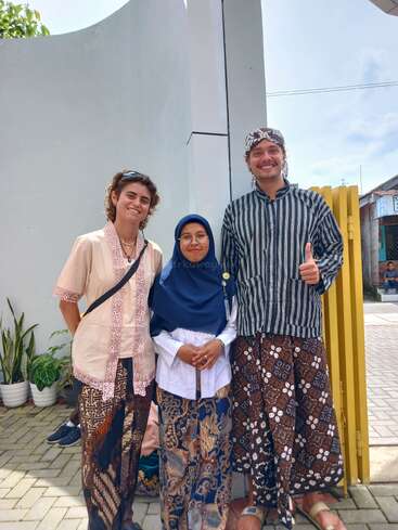 Three people wearing traditional Indonesian clothes pose together outdoors, smiling happily. Two are foreigners; all wear batik. Potted plants and a modern building are visible behind them.