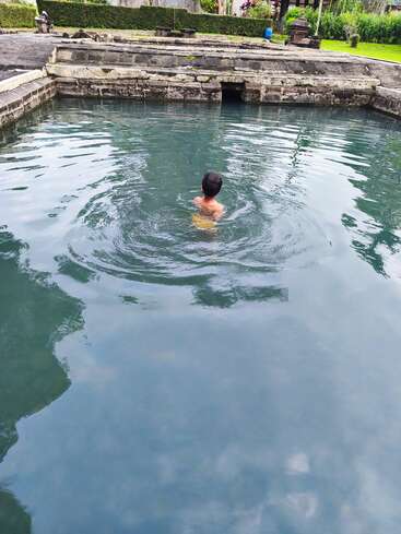 Un niño nada solo en una piscina de piedra cristalina y tranquila, rodeada de antiguos muros de piedra y una exuberante vegetación, lo que crea un ambiente apacible y sereno.