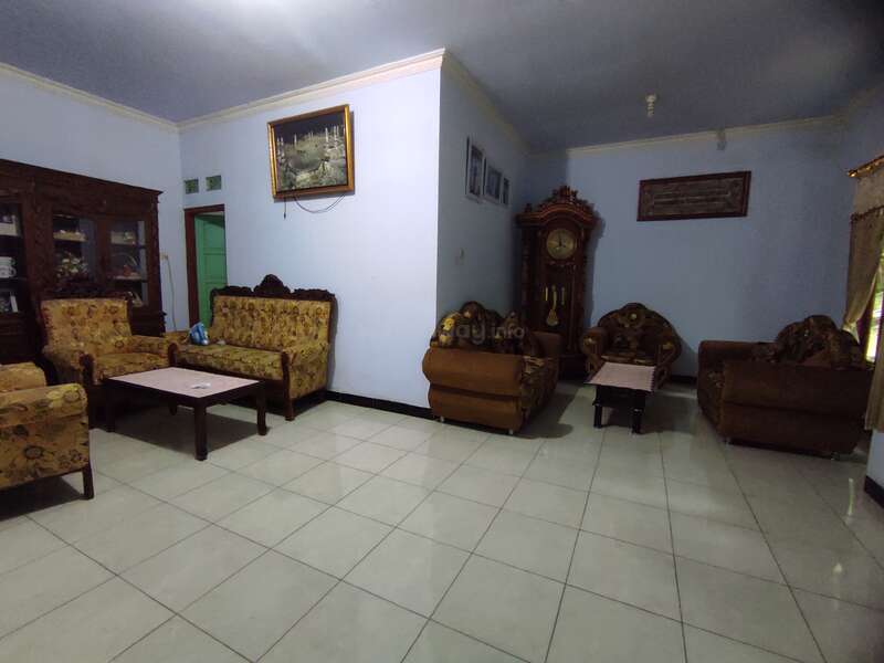 This living room features vintage sofas, a large grandfather clock, a glass-fronted cabinet, framed wall art, tiled flooring, and pale blue walls with yellow curtains.