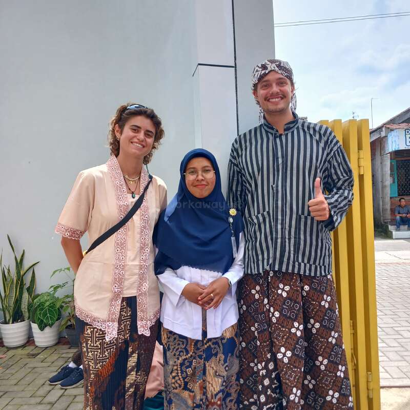 Tres personas vestidas con ropas tradicionales indonesias posan juntas al aire libre, sonriendo alegremente. Dos son extranjeros; todos llevan batik. Detrás de ellos se ven plantas en macetas y un edificio moderno.
