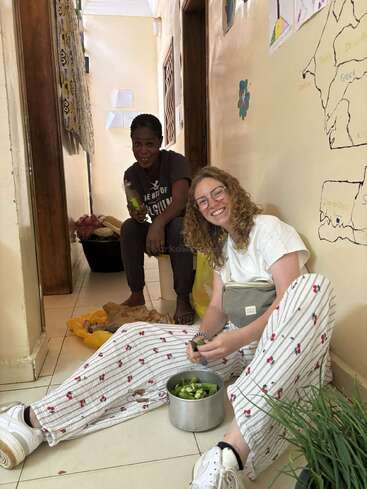Deux femmes préparent joyeusement des légumes dans un couloir. L'une est assise par terre et sourit, tandis que l'autre est assise derrière, un couteau à la main.