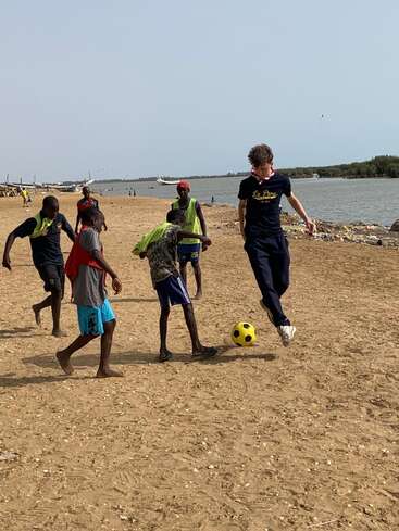 Un groupe d'enfants joue au football sur une plage de sable près de l'eau, s'amusant ensemble sous un ciel clair, mettant en valeur le plaisir et le travail d'équipe.
