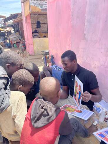 A man sits beside a pink wall, showing illustrated educational posters to a group of attentive children gathered around him in an outdoor, urban market setting.