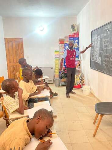 In a bright classroom, an instructor teaches young boys seated at desks. The boys attentively write notes while the teacher points to the blackboard with lessons.