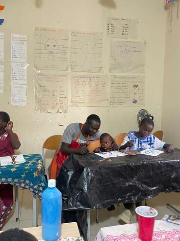 A teacher helps two young children with painting activities in a classroom. Educational posters in French decorate the wall, creating a lively, creative learning environment.