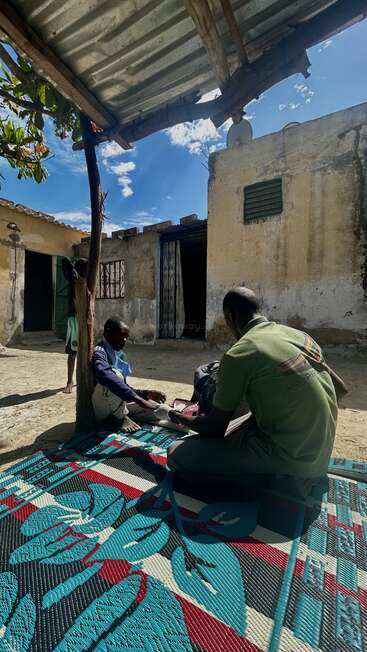 Three people are outdoors under a tin roof in a modest compound. Two sit on a colorful mat, one receiving medical attention, while another stands nearby.
