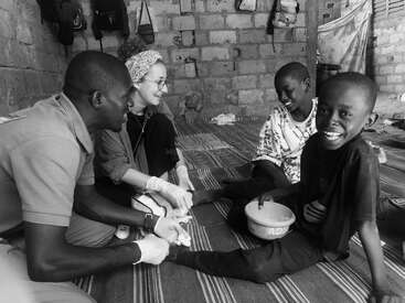Two adults, wearing gloves, tend to two smiling children’s feet in a modest room. All appear happy, suggesting a caring interaction, possibly medical or humanitarian aid.