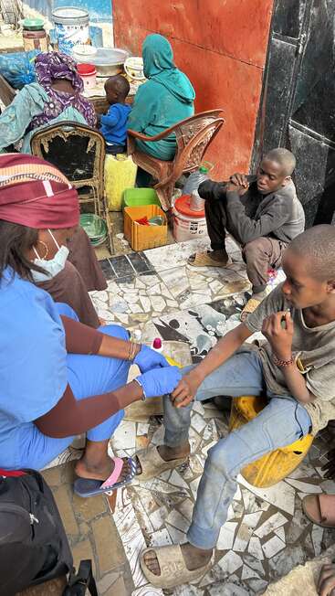 A healthcare worker in blue scrubs treats a young boy’s hand outdoors. Several people sit nearby, surrounded by buckets and food containers on mosaic tiles.