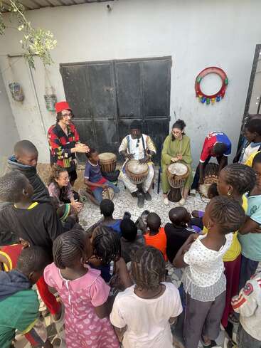 A group of children gathers in a courtyard, enthusiastically watching and listening to three adults playing traditional drums, sharing music and cultural learning together.