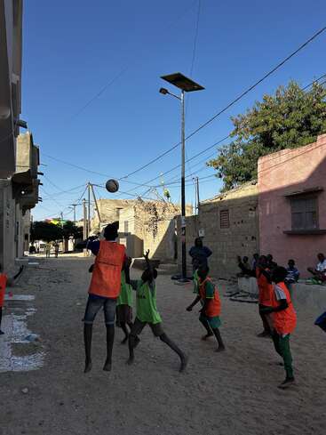 A group of children wearing orange and green vests play a lively game of basketball on a sandy street, surrounded by buildings, under a clear blue sky.