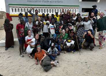 A joyful group of children and adults pose together outside, smiling and celebrating in front of a building marked “Association d’aide aux enfants” in French.