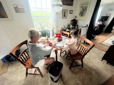 Uma mulher e uma criança sentam-se em uma mesa de madeira em uma cozinha aconchegante e iluminada pelo sol, preparando a comida juntas, cercadas por plantas, arte e decoração caseira.
