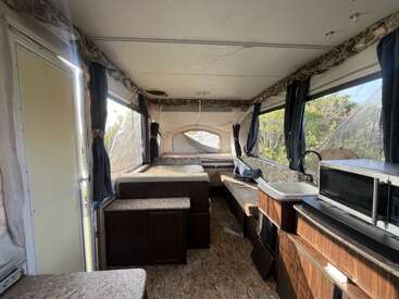 Esta imagem mostra o interior de um trailer pop-up. Ele tem camas, uma cozinha compacta com pia e micro-ondas, janelas com cortinas e armários marrons.