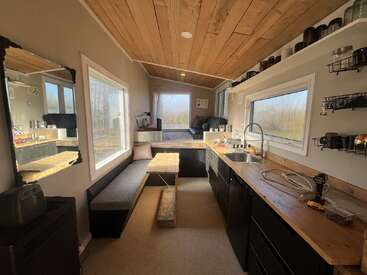 Dieses Bild zeigt ein gemütliches, modernes kleines Haus mit Holzdecke, effizienter Küche, Sitzbank, großen Fenstern, offenen Regalen und natürlichem Licht, das eine warme Atmosphäre schafft.