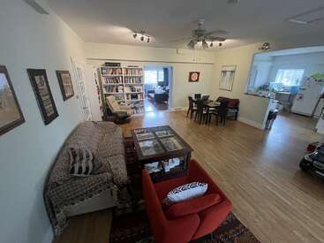 Dieser geräumige Wohnbereich verfügt über ein gemustertes Sofa, einen roten Sessel, einen Couchtisch, ein Essgeschirr, ein Bücherregal, einen Holzfußboden, einen Deckenventilator, eine Küche und viel Tageslicht.