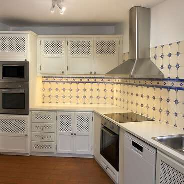 Cette cuisine est équipée d'armoires blanches aux portes ajourées, de carreaux à motifs bleus et blancs, d'appareils électroménagers modernes, d'une hotte en acier inoxydable, d'une plaque à induction et d'un sol en terre cuite.