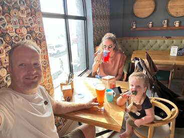 Eine dreiköpfige Familie sitzt an einem sonnigen Restauranttisch. Der Vater macht ein Selfie, die Mutter trinkt etwas, und das Baby hat einen Schnuller und ein Spielzeug.