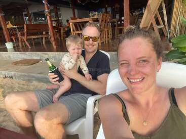 Eine lächelnde Frau zwinkert der Kamera zu, während sie ein Selfie macht; neben ihr sitzt ein Mann mit einem Baby und einem Getränk in der Hand und entspannt sich im Außenbereich eines Restaurants.