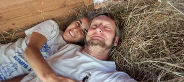 Un couple heureux est allongé sur un lit de paille à l'intérieur d'une structure en bois, souriant chaleureusement à la caméra, rayonnant d'amour, de confort et d'intimité dans un décor rustique.