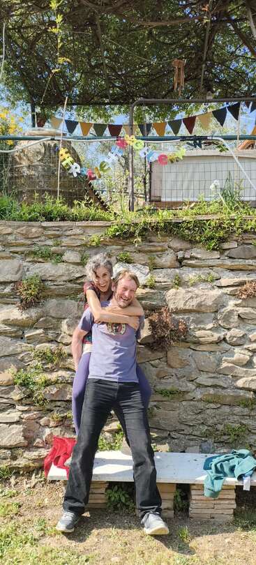 Un homme souriant porte une femme sur son dos à l'extérieur, sous des décorations festives, dans un jardin ensoleillé avec des murs en pierre et une végétation luxuriante. Un moment joyeux.