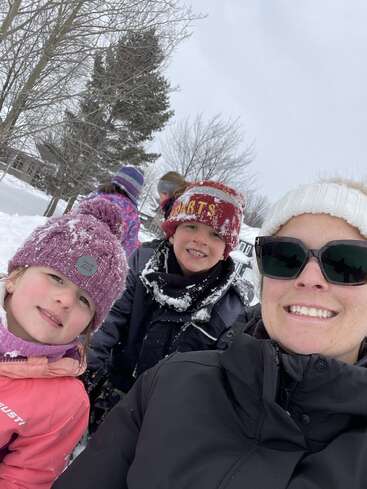 Eine Frau und drei in Winterkleidung gekleidete Kinder lächeln im Freien. Der Schnee bedeckt den Boden und die Bäume und sorgt für eine fröhliche, verschneite Tagesstimmung mit Familienspaß.