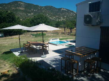 A sunlit outdoor patio features wooden tables, chairs, umbrellas, a small pool, bar stools, lush greenery, and scenic mountains in the background under a clear sky.