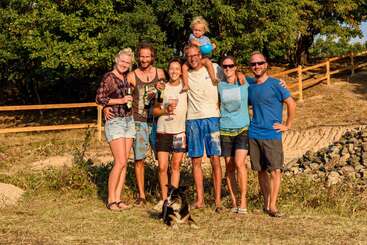 The image depicts a group of seven people, including a child on an adult\'s shoulders, standing in a grassy area with trees in the background, posing for a photo.
