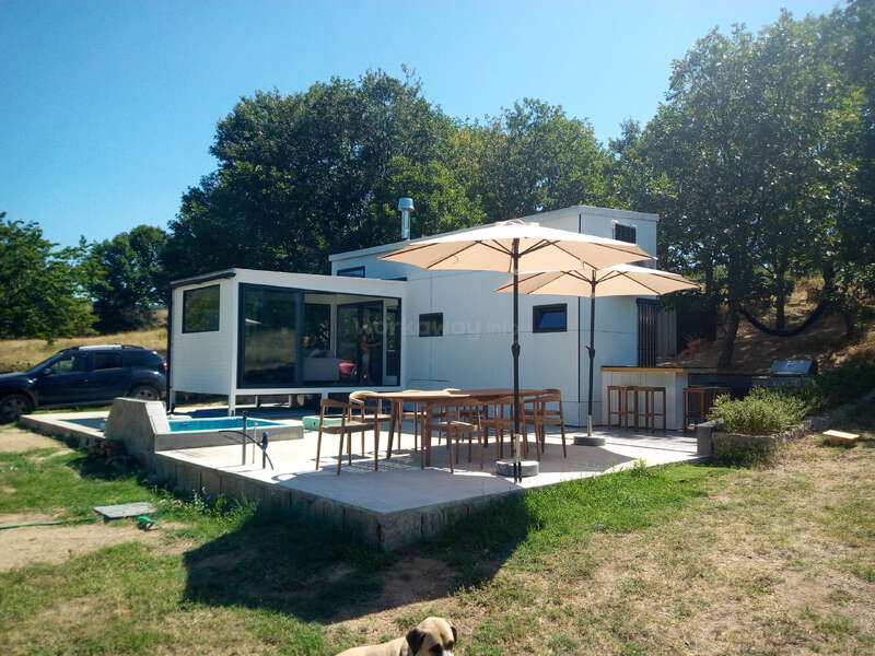A modern tiny house with a patio featuring tables and umbrellas. Surrounded by trees, a car is parked nearby. There's a dog resting in the foreground.