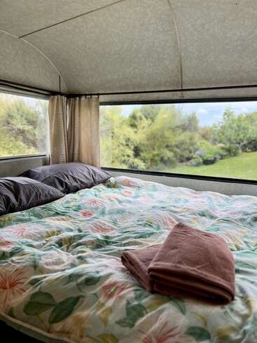 Una acogedora cama con ropa de cama floral y toallas dobladas se encuentra dentro de una caravana con grandes ventanas, que ofrecen una hermosa vista de la vegetación y la naturaleza del exterior.