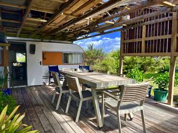 Una acogedora zona de descanso al aire libre con una mesa larga y sillas se encuentra bajo una pérgola de madera junto a una caravana retro, rodeada de exuberante vegetación y luz solar.