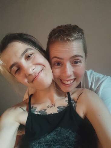 Deux personnes sourient joyeusement, proches l'une de l'autre, en prenant un selfie. La personne à l'avant a des tatouages visibles et toutes deux semblent heureuses, détendues et apprécient la compagnie l'une de l'autre.