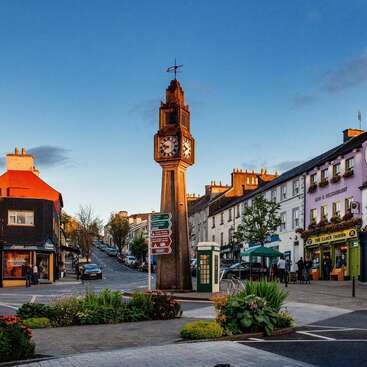 A quaint town square features a tall clock tower, colorful buildings, shops, a tavern, signposts, lush flowerbeds, and people enjoying a peaceful, vibrant evening atmosphere.