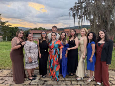 A group of ten women and one man, dressed formally, stand together outdoors on stone pavement with greenery, trees, and cloudy sky in the background, smiling.