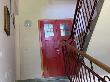 Une double porte rouge avec des panneaux en verre se trouve en haut d'un escalier, avec une rampe en métal et des murs beiges ornés de photos encadrées.
