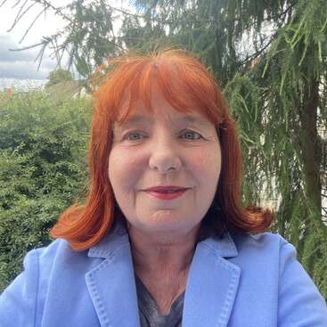 A woman with red hair smiles at the camera, wearing a light blue blazer. Green trees and foliage fill the background, with a cloudy sky above.