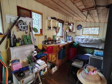 Un intérieur de cabine confortable et encombré avec des murs en bois, des étagères, des livres, un lit, des ustensiles de cuisine, des fleurs, un chapeau de paille, des œuvres d'art et la lumière naturelle provenant de plusieurs fenêtres.