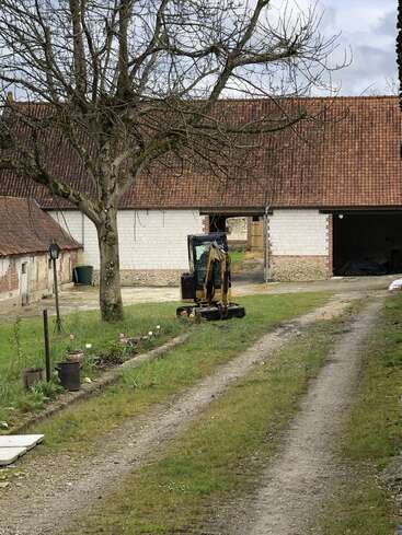 Eine unbefestigte Auffahrt führt zu einer rustikalen Scheune mit rotem Ziegeldach. Ein kleiner Bagger steht neben einem kahlen Baum auf dem grasbewachsenen Hof.