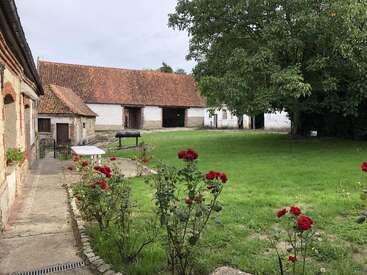 Ein friedlicher ländlicher Innenhof mit grünem Gras, blühenden roten Rosen, alten Backsteinhäusern, einem großen schattenspendenden Baum, einem Grill und einer ruhigen, beschaulichen Atmosphäre.