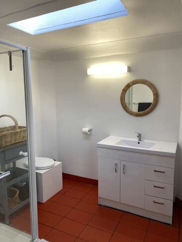 Esta é uma casa de banho moderna com piso de azulejos vermelhos, um lavatório branco, um espelho redondo, uma claraboia, um vaso sanitário, um box de vidro, uma cesta de arrumação e um aplique de parede bem iluminado.