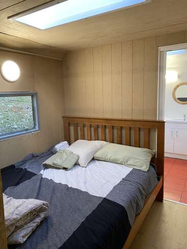 Um quarto aconchegante com paredes revestidas de painéis de madeira, uma cama de madeira e roupa de cama cinza. A luz natural entra pelas claraboias e por uma janela. Dá para ver o banheiro, com piso de azulejos vermelhos.