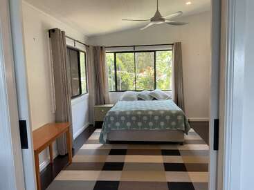 Esse quarto tem uma cama de casal com roupa de cama estampada, janelas grandes com cortinas, ventilador de teto, mesas laterais e um tapete xadrez sobre piso escuro.