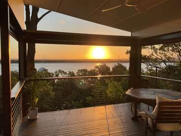 Uma varanda tranquila tem vista para um pôr do sol incrível sobre águas calmas, com árvores abaixo, veleiros ao longe, assentos aconchegantes e a luz do sol quente e dourada iluminando tudo.