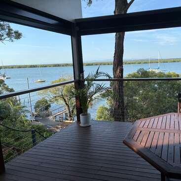 Um deck de madeira tem vista para um lago tranquilo com veleiros, cercado por árvores frondosas. Uma mesa, cadeira, vaso de planta e xícara de café criam um refúgio tranquilo.