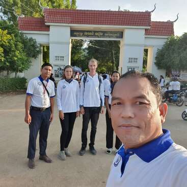 Das Bild zeigt eine Gruppe von Menschen vor einem Gebäude mit einem roten Dach, umgeben von Bäumen und Motorrädern, wobei eine Person im Vordergrund ein Selfie macht.