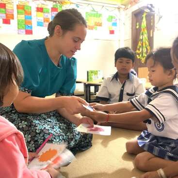 Una mujer con un top azul entrega un papel a un niño en un aula, rodeada de otros niños y de coloridos adornos en las paredes.