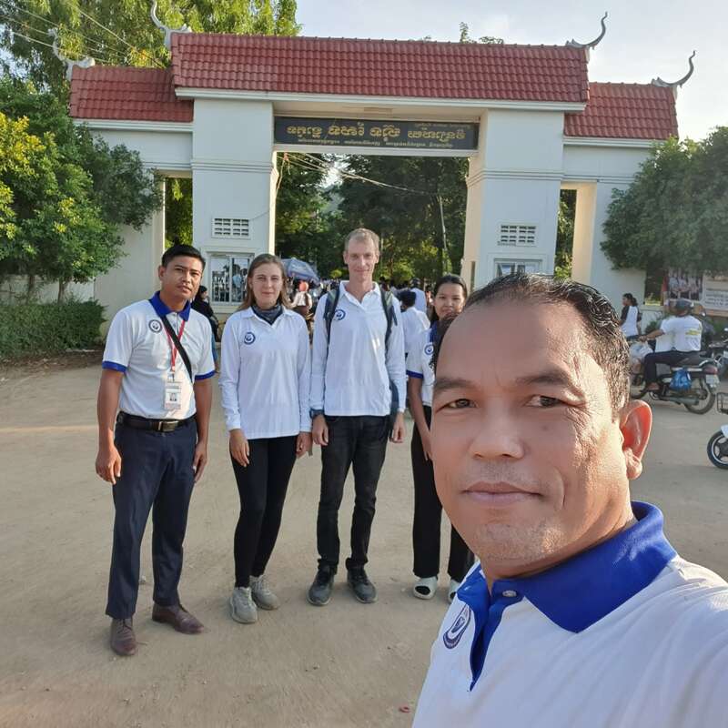 La imagen muestra a un grupo de personas delante de un edificio con tejado rojo, rodeado de árboles y motocicletas, con una persona tomándose un selfie en primer plano.