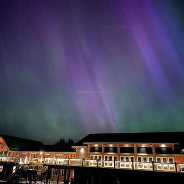 A cozy, illuminated lodge sits beneath a stunning night sky filled with vibrant purple and green aurora borealis lights, creating a magical, tranquil, and mesmerizing atmosphere.