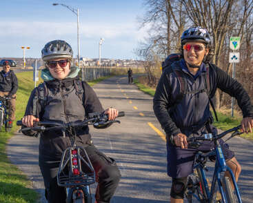 Dois ciclistas, usando capacetes e óculos escuros, sorriem enquanto pedalam em uma trilha pavimentada ensolarada. Você pode ver árvores sem folhas, a paisagem urbana e outro ciclista ao fundo.