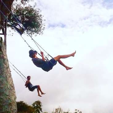 The image depicts two individuals on a swing suspended from a tree, with one person in the foreground and another in the background, set against a cloudy sky.